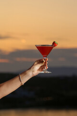 Woman holding a glass of strawberry daiquiri cocktail with beautiful vibrant sunset behind