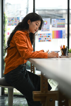 Concentrated young creative woman sitting in bright office and working on design project.
