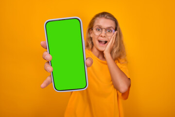 Young shocked attractive Caucasian woman holds mobile phone with green screen and opens mouth in...