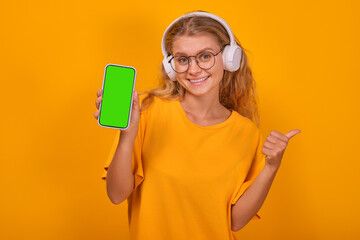 Young cheerful casual Caucasian woman teen in headphones showing smartphone with green screen and thumb up recommending you to place commercial offer on phone display stands on orange background.