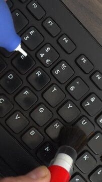 Vertical video social media format – Closeup overhead shot of hands using a blower and brush to clean the keys of a dirty and dusty computer keyboard.