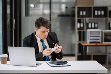 Happy young man looking at cellphone screen, reading message, or email, communicating distantly with client or playing games from computer work in modern office.