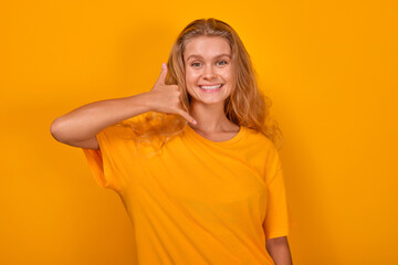 Young cheerful beauty Caucasian woman in casual t-shirt makes call me gesture and smiling looks at screen offering to exchange phone numbers to be in touch stands in orange studio.