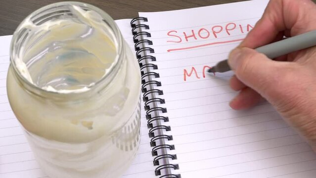 Closeup of a hand using a felt pen to write mayo in a lined notebook, under the heading of shopping, next to an empty glass jar of mayonnaise.