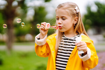Cheerful little girl in a yellow raincoat blows soap bubbles, plays on a walk in the park in...