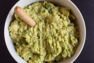 A macro shot of a delicious homemade guacamole dip in a round bowl, garnished with a crunchy whole grain breadstick. Traditional food