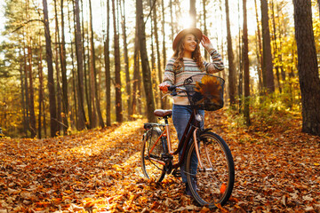 Smiling young woman in a hat and a stylish sweater with a bicycle walks and enjoys the autumn...