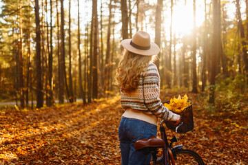 Smiling young woman in a hat and a stylish sweater with a bicycle walks and enjoys the autumn weather in the forest, among the yellow leaves at sunset.