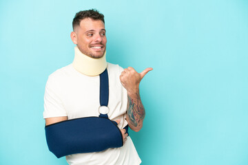 Young caucasian man wearing a sling  and neck lace isolated on blue background pointing to the side...