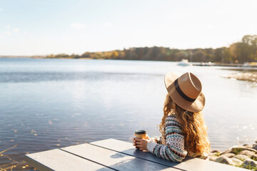 Young tourist woman in a hat drinking coffee and enjoying the sunny weather on a bench by the lake. Concept relax, travel, nature. Active lifestyle