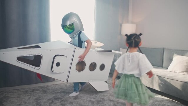 Chinese kids play in the living room of the house, a boy in an astronaut helmet plays with a cardboard space shuttle, a cute girl sister runs with brother around the room.