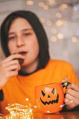 Portrait of candid authentic zoomer boy posing with jack lantern mug and sweets at halloween party