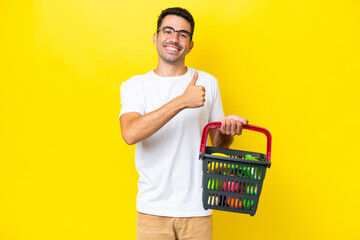 Young handsome man holding a shopping basket full of food over isolated yellow background giving a...