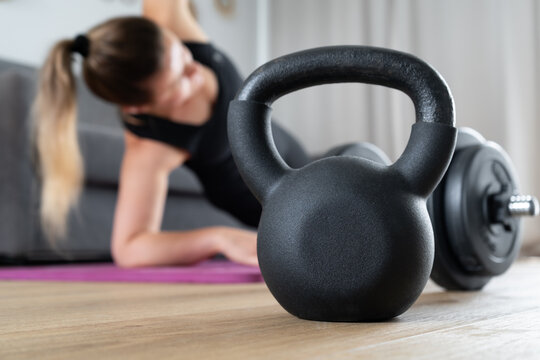 Woman exercising at home in the living room on yoga mat. House fitness workout, sport training concept. Focus on gym exercise equipment, kettlebell, dumbbell barbell. Female doing pilates, stretching.
