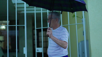Senior man opening umbrella to protect from rain, steps out in the street from home. Elderly person...