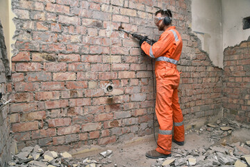 Worker standing removing old plaster from red brick wall by hammer drill, wearing gloves, boots,...