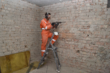 Construction worker removing old cement from brick wall indoor, standing on the ladder using hammer...