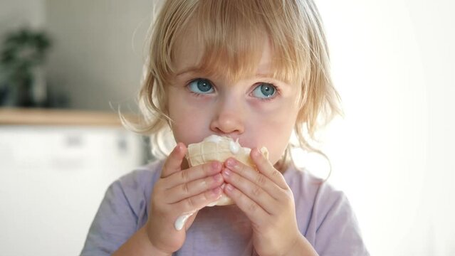 Baby girl enjoying ice cream. Pretty little toddler eating an ice-cream indoors, at home. Dining room background. Small child eats plombir and cream messy on her mouth. Cute kid with tasty sweet food.