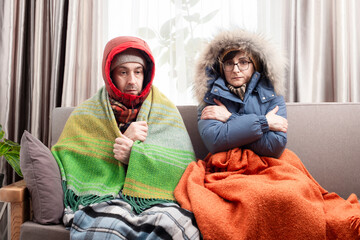 Sad middle age couple in warm clothes getting cold on the sofa in their living room