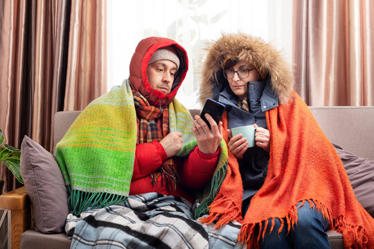 Middle age family in parkas drinking coffee on the sofa while using smartphone in their cold living room