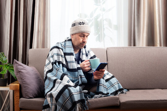 Middle age male model in hat and plaid hold coffee cup sitting on the sofa while using his smartphone