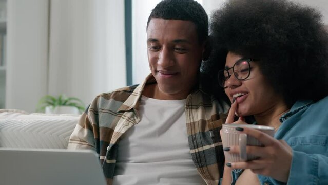 African American happy family couple man woman girlfriend boyfriend husband wife with cup of tea coffee looking at laptop shopping online store shop at home watching movie film with computer talking