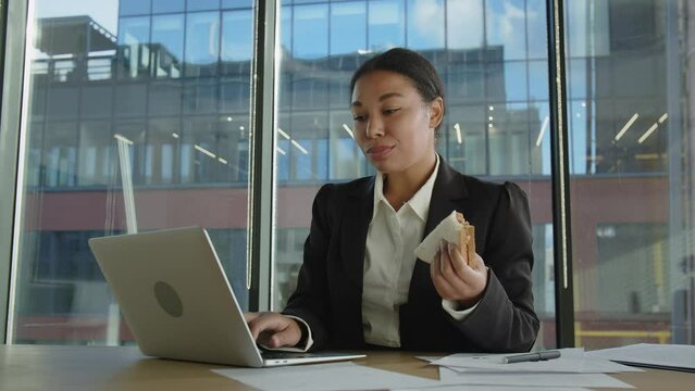 Young afro-american woman type on pc something with one hand and eating with another. Black woman in office eating sandwich for a breakfast because has no time. Stressful work and eating unhealthy