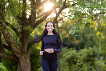Young beautiful woman running in the park, portrait of a Latin American woman jogging and doing active exercises, sportswoman smiling and looking at the camera among the trees.