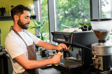bearded barista cleaning coffee machine nozzle with rag while working in coffee shop