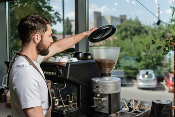 side view of barista in apron using coffee grinder while working in coffee shop on background