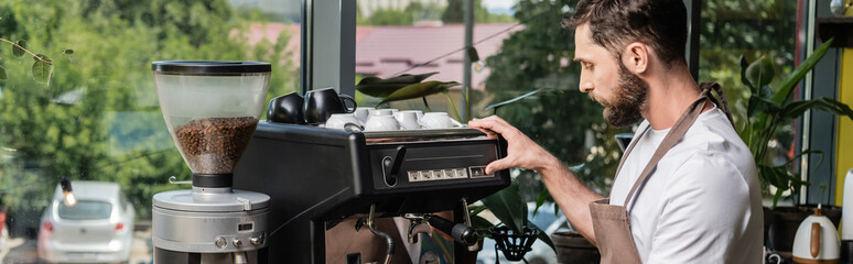 side view of bearded barista in apron using coffee machine while working in coffee shop, banner