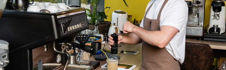 cropped view of barista in apron grinding coffee near digital tablet and cold coffee in cafe, banner