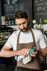 Fototapeta na wymiar bearded and focused barista in apron cleaning cup with towel while working in coffee shop