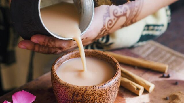 Indian woman hands pouring masala chai tea from Cezve into wooden cup.