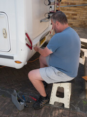 A male mechanic sitting down in shorts works on the rear lights of a vehicle with a...