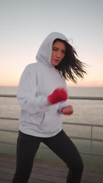 Kickboxer woman dressed hoodie warming up arm and leg muscles before training. Athletic middle age female boxer boxing workout outdoor on pier. Vertical video