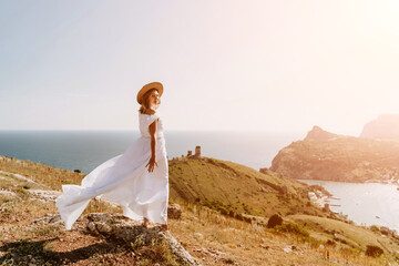 Fototapeta na wymiar Happy woman in a white dress and hat stands on a rocky cliff above the sea, with the beautiful silhouette of hills in thick fog in the background.