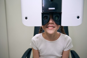 Cheerful girl sits in front of special apparatus for diagnosing vision