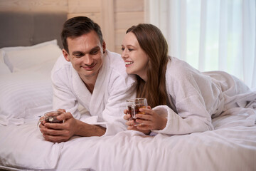 Obraz na płótnie Canvas Smiling couple wearing bathrobes and laying in bed with teacups