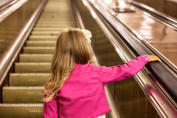 Rear view of little girl child 5-6 year old wear pink jacket in subway metro, going up escalator. Kid passenger in underground subway, from behind. Public urban transport concept. Copy ad text space