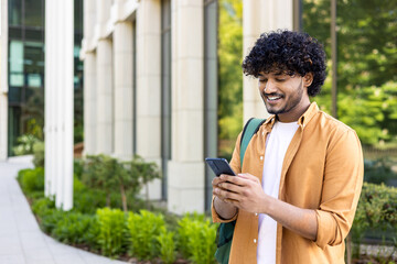 Young smiling Indian male student, freelancer, tourist standing outside and holding phone. Writes a message, looks for a way, calls