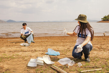 Portrait beautiful woman sitting with thumbs up holding plastic bottle together with male volunteers collecting plastic waste and putting on a garbage bag natural fresh water source by the beach.