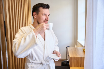 Smiling man in soft bathrobe enjoying morning drinking coffee