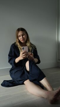 a girl in a blue dress sits on the floor against the background of a white wall, talks on the phone via video call, then looks at the camera, waves her hands and laughs. vertical video