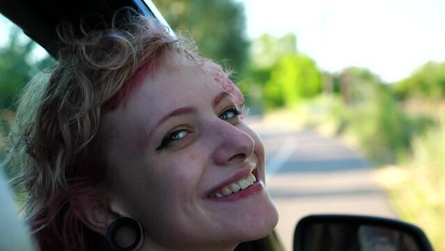 Close up to head of happy punk girl with ear tunnels leaning out of auto window and looking into camera. Smiling hippie woman enjoying car journey at summer day. Adventure and travel concept. Slow mo