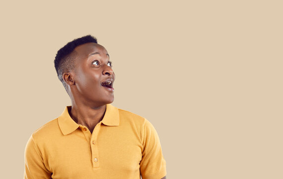 Young positive WOW African American man looks up with enthusiastic emotions when he sees unexpected promotional offer or surprise with huge discounts for shopping, stands in beige studio. Copy space