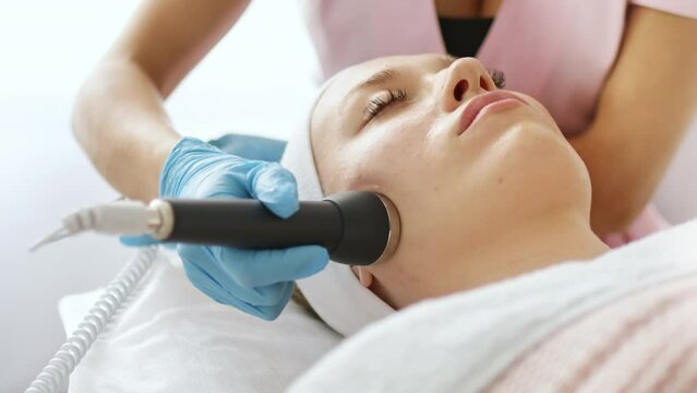 Beautician makes a procedure on the face on the cheekbones and nasolabial folds with microcurrent therapy device. Hardware cosmetology in clinic salon for client.	