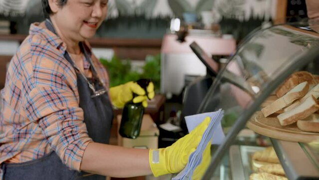 Asian mothers are still healthy, using cloth wipe glass cabinet in daughter bakery, spray glass cleaner down, use thin cloth wipe it clean, after opening shop all day maintain cleanliness in store.