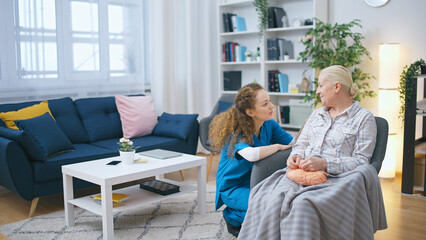 An old woman sits in an armchair, knitting, while a nurse visits to check up on her