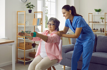Elderly active woman exercising with blue elastic band. Attentive female physiotherapist or...